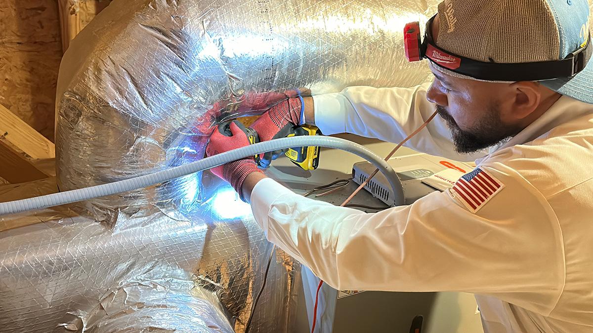 A technician in a white uniform repairs the insulated ductwork near the furnace, using a tool and wearing a headlamp, gloves, and a cap.
