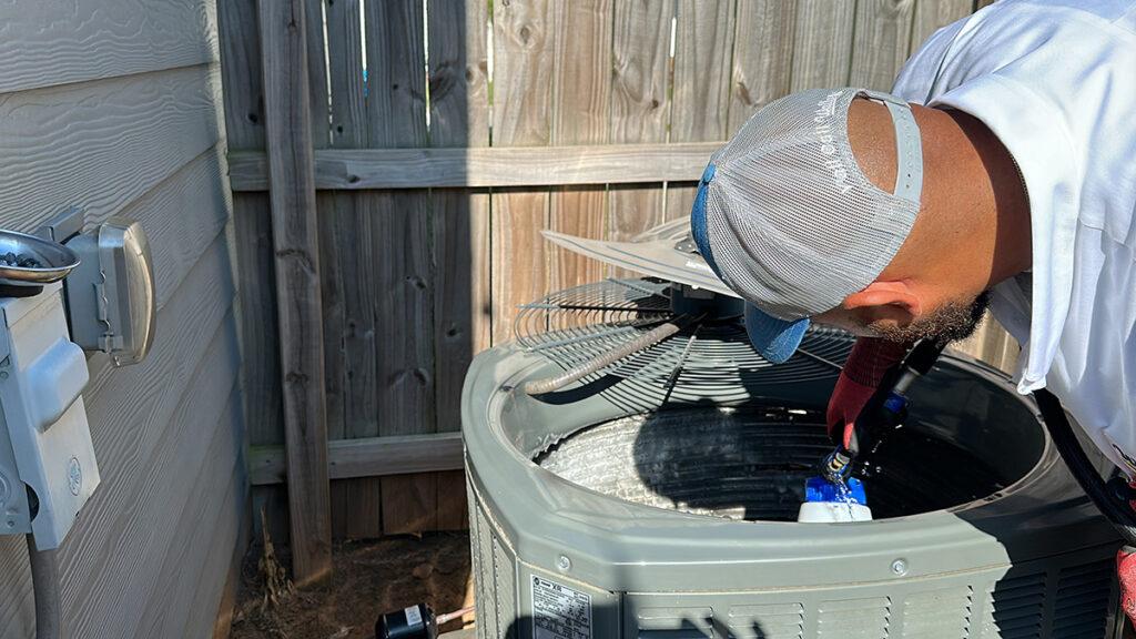 Wearing a cap and gloves, the man diligently repairs an outdoor air conditioning unit next to a wooden fence and wall, ensuring it runs smoothly.