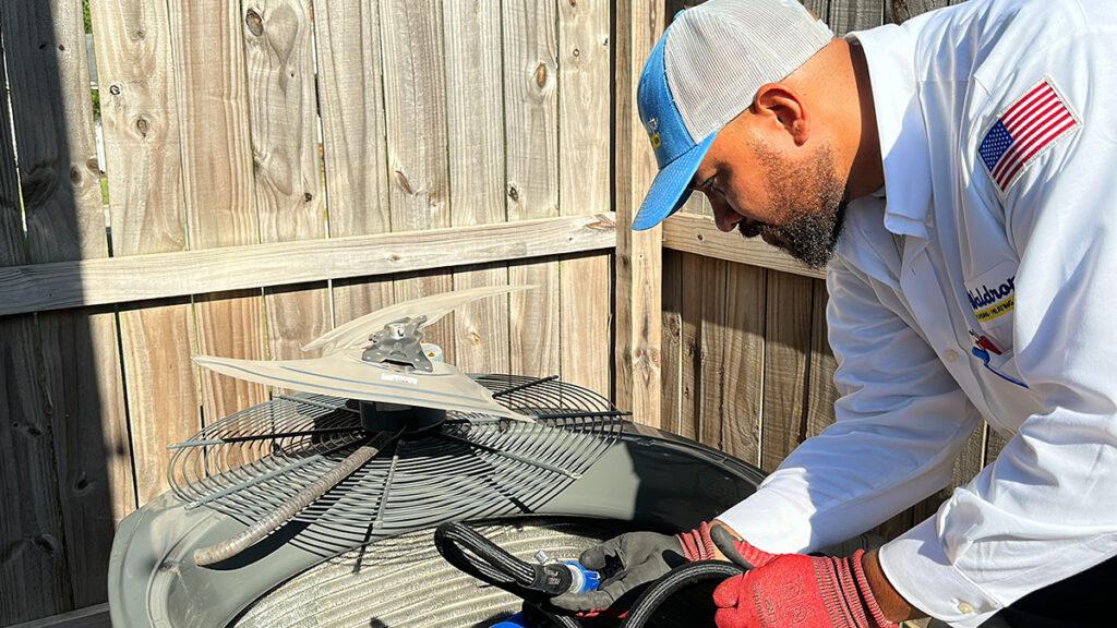 A technician in a white uniform and cap, wearing red gloves, adjusts an outdoor air conditioning unit near a wooden fence on a sunny day, working to ensure seamless operation like a seasoned plumber.