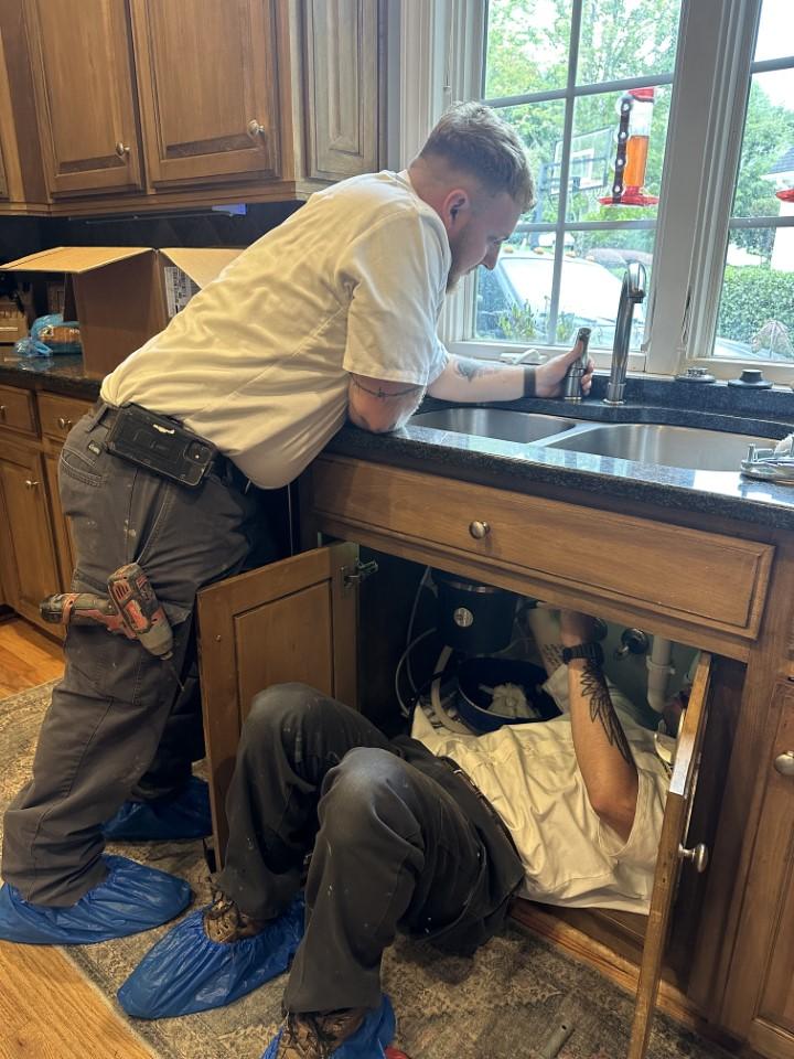 Two plumbers, both wearing white shirts and blue shoe covers, work on a kitchen sink. One lies under the sink with tools, while the other stands by the counter.