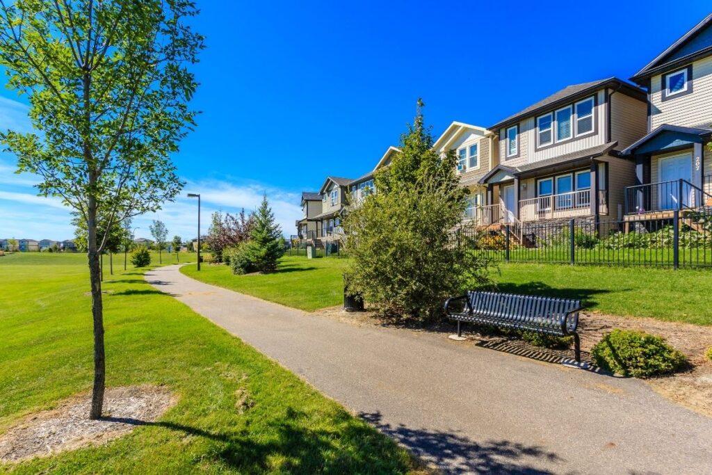 Wally walks a paved sidewalk by grassy fields and modern houses, passing benches and trees under clear blue skies toward Anderson.