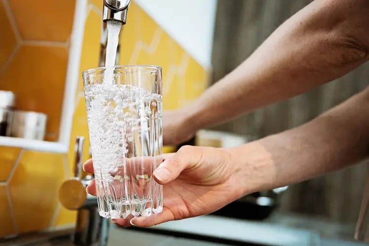 A person fills a clear glass with water from a running kitchen faucet connected to a whole-house filter. Yellow tile backsplash shows.