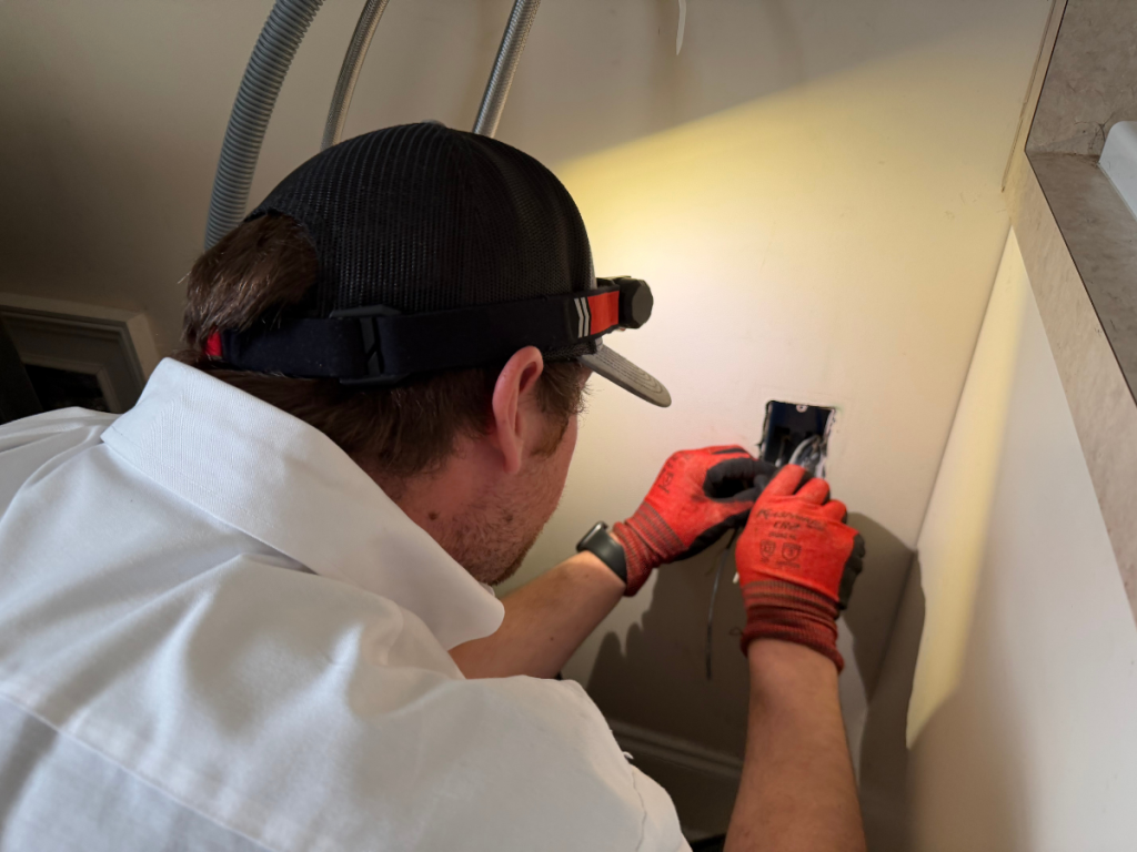 Electrician wearing a headlamp and orange gloves installing an outlet in a bathroom wall near a sink/bathtub area.