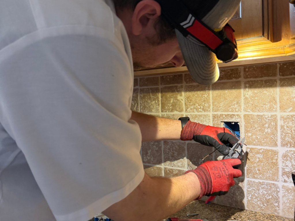 Worker wearing gloves and a cap installs or repairs an electrical outlet in a tiled kitchen backsplash. Wires and a blue outlet box are visible behind the opening.