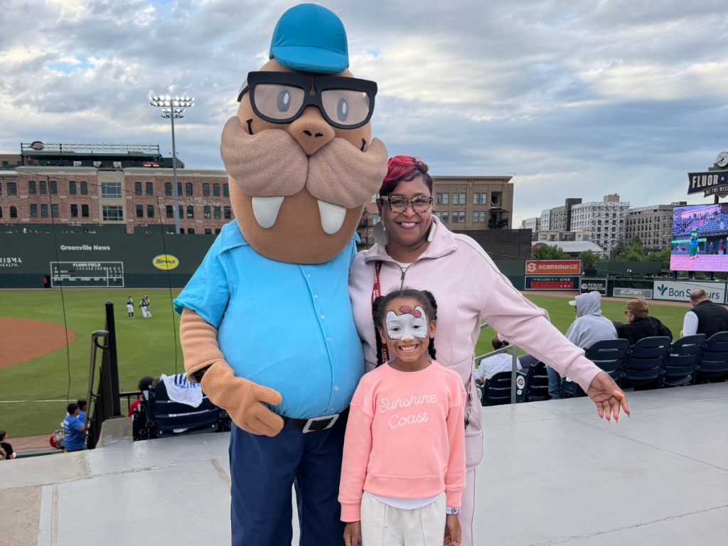 Girl with face paint posing with a smiling woman and a giant cartoon beaver mascot in a blue shirt at a baseball stadium.