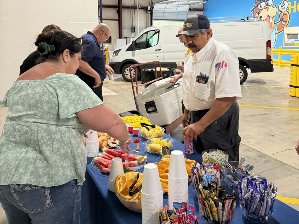 People at a food table in a warehouse: a man in a white shirt and cap hands out fruit and snacks to a woman in a green floral blouse.