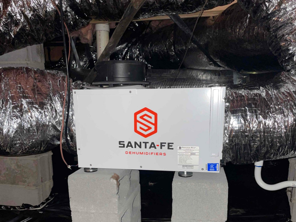 White Santa-Fe dehumidifier installed in a crawl space, sitting on concrete blocks among ductwork and insulation, with a drain hose nearby
