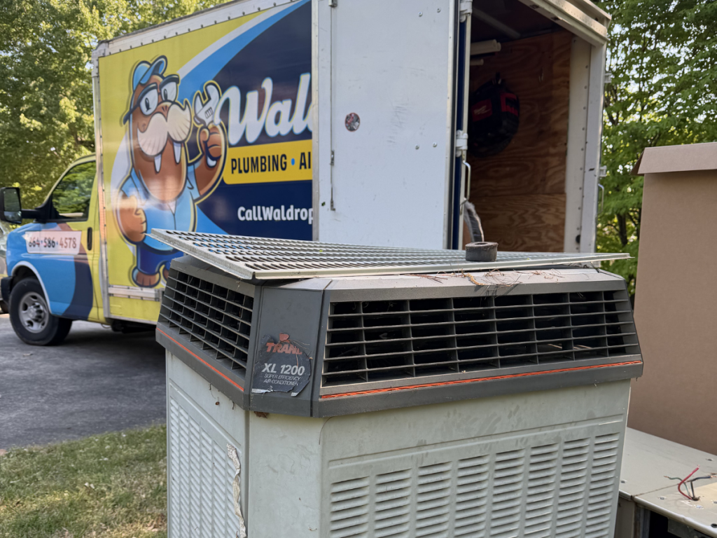 Gray outdoor HVAC a/c unit with a metal top grate in a driveway; a yellow-blue plumbing truck with a cartoon beaver logo is parked behind it, open cargo area visible.