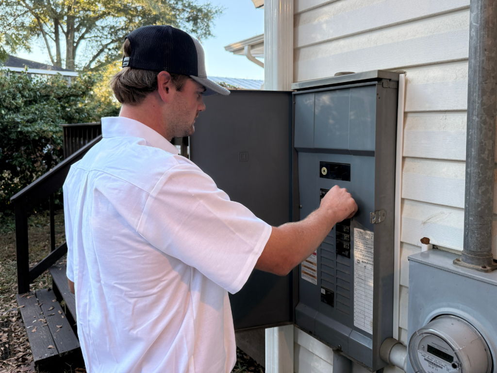Man in a white shirt and baseball cap operating an outdoor electrical panel mounted on a house exterior.