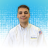 Portrait of a smiling technician in a white button-down shirt with a Waldrop Plumbing, Heating & Air patch on the chest and an American flag patch on the sleeve, against a blue dotted background.
