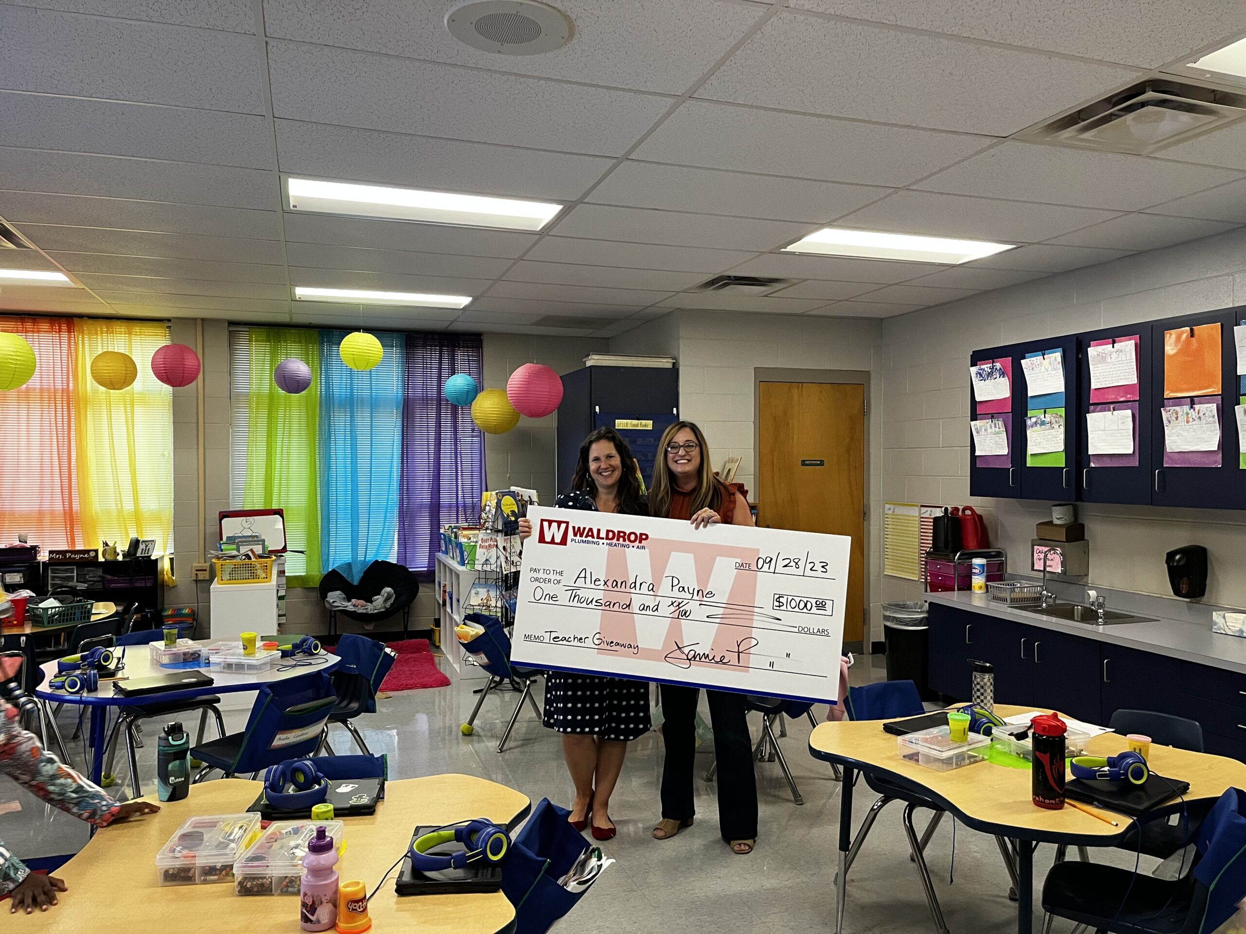 Two women in a classroom hold a large ceremonial check for $5,000.0 with colorful rainbow curtains in the background, celebrating a giveaway event in a school.