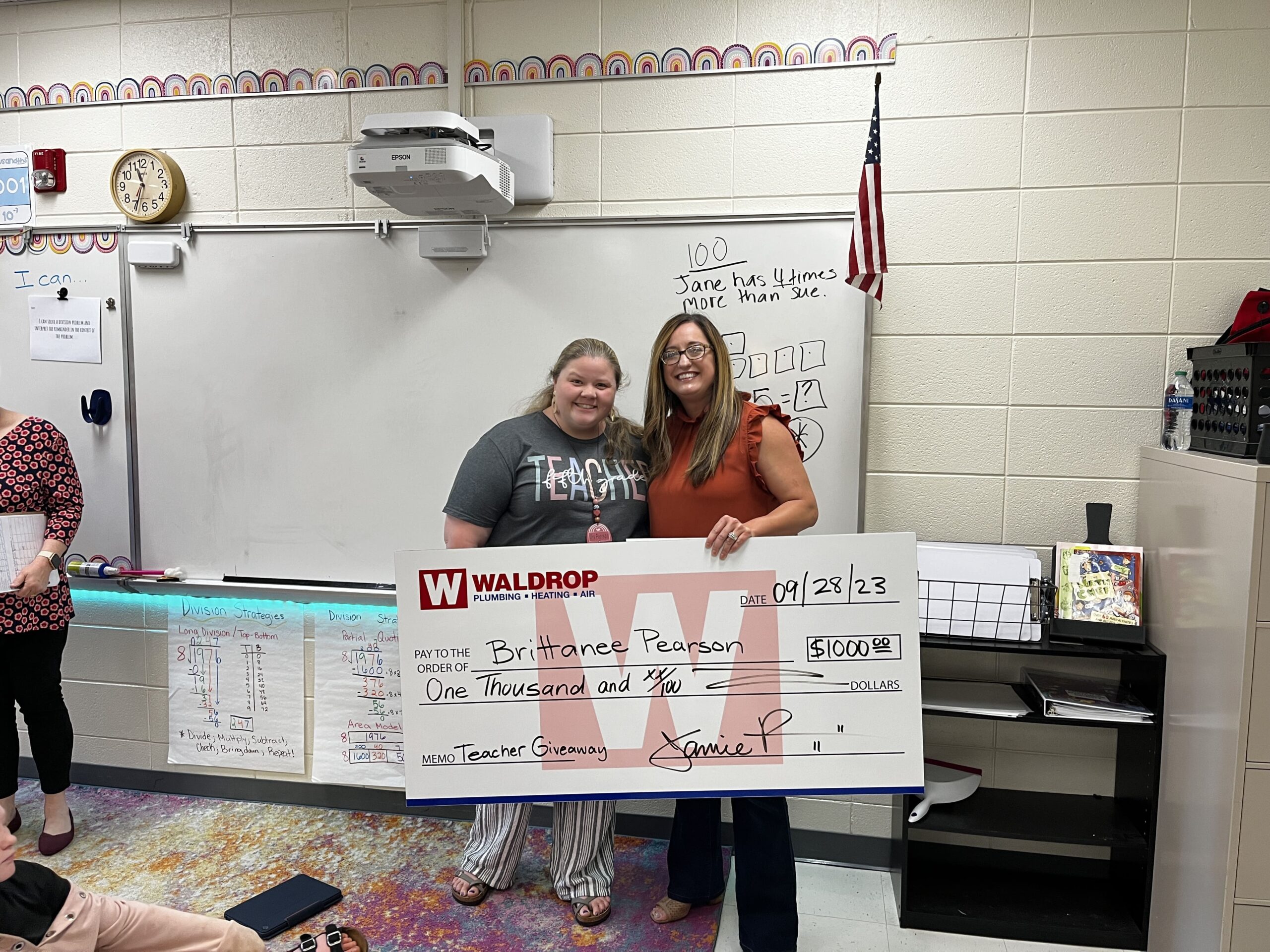 Two smiling women hold a large ceremonial check in a classroom, with a whiteboard, projector, and American flag in the background.
