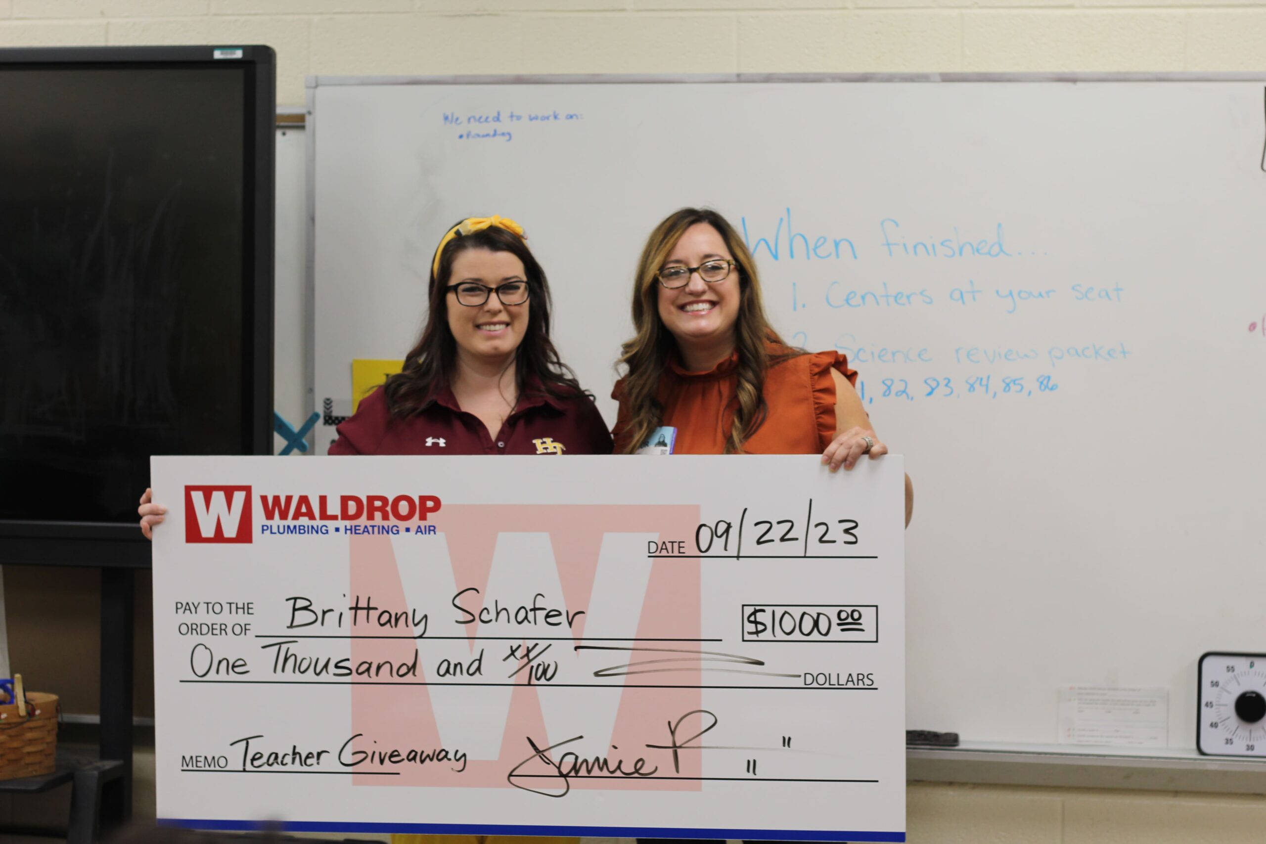 Two smiling women stand behind a large ceremonial check from WalDROP Plumbing, Heating & Air, posing for a teacher giveaway event.