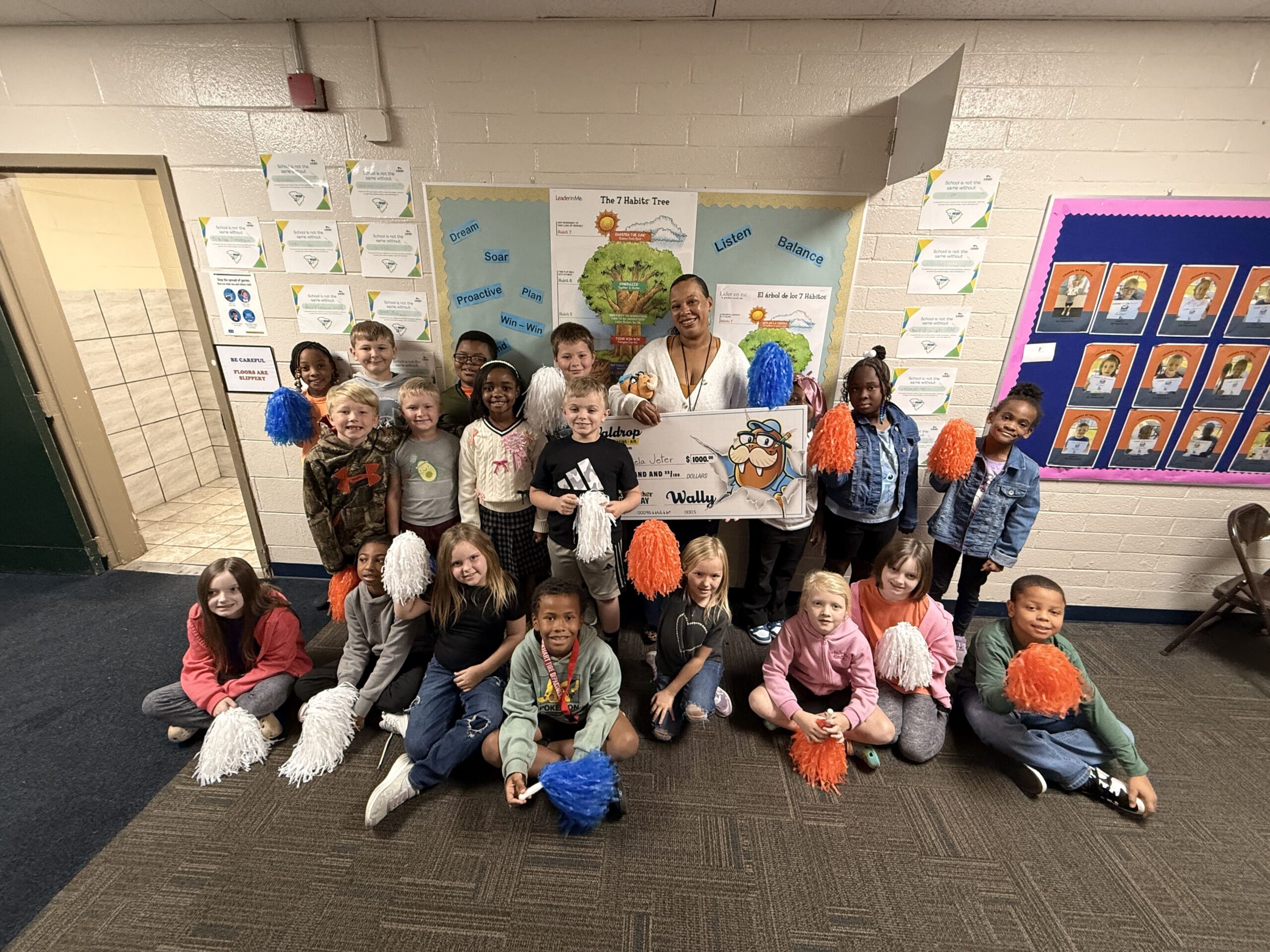 Group of children and a teacher posing in a school hallway with colorful pom-poms and classroom posters behind them.