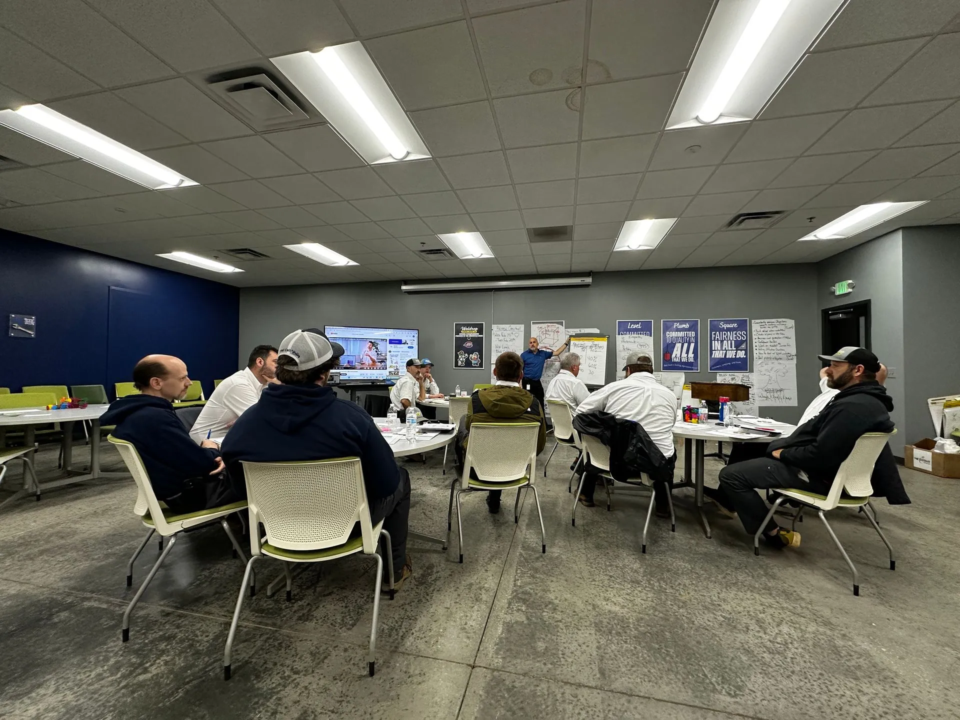 Group of people seated around round tables in a classroom, watching a presenter at the front with a TV screen and posters on the wall.