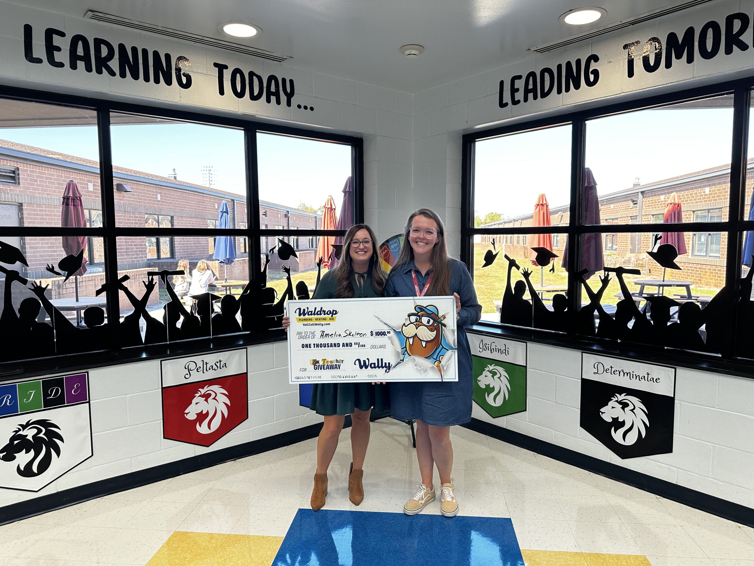 Two smiling girls hold a large ceremonial check in a school hallway with student silhouettes on the display boards and banners reading 'Learning today... Leading tomorrow' above them via windows. (informative)