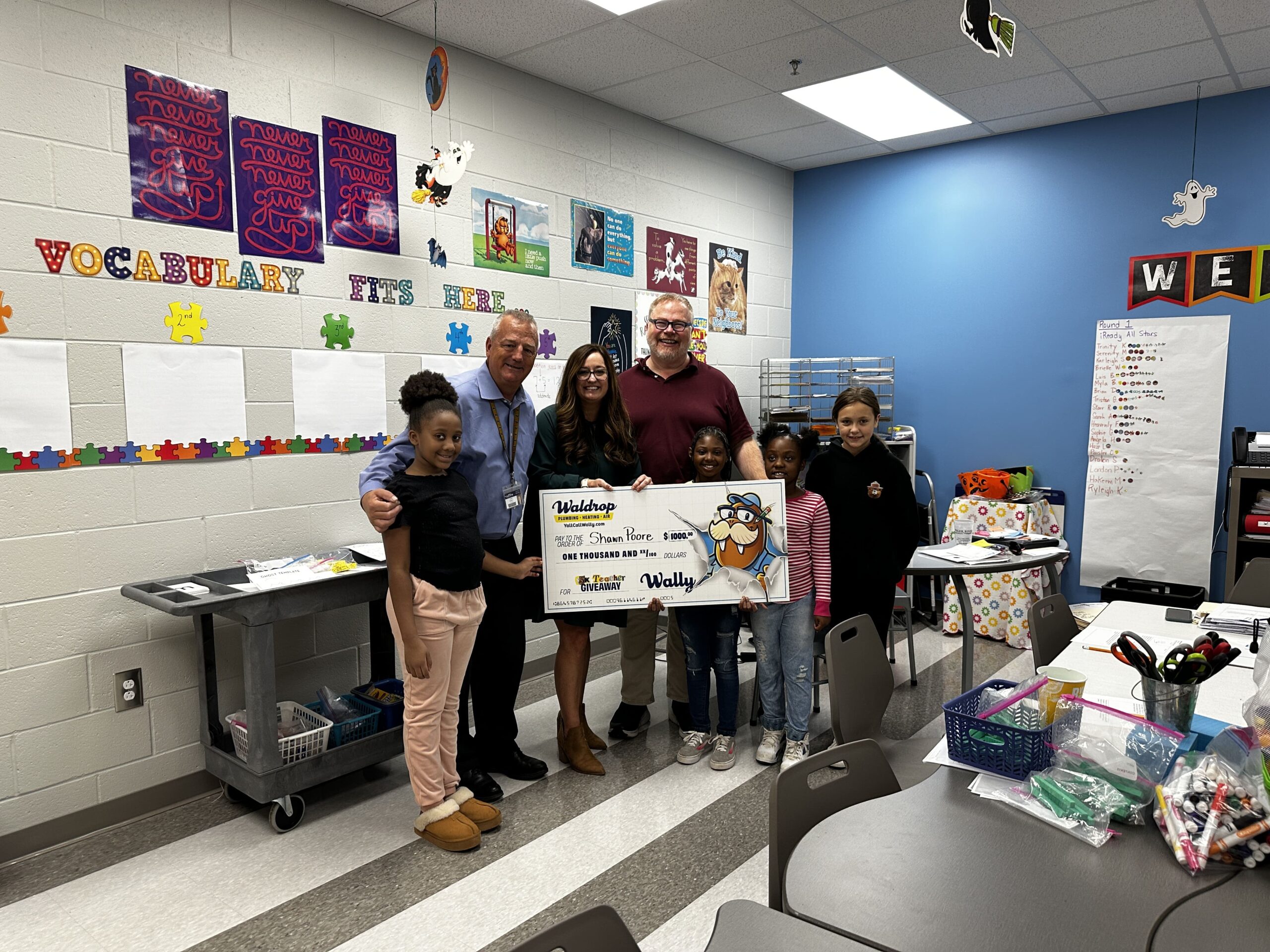 Group of adults and children posing with a large cartoon-style check in a classroom, with colorful vocabulary posters and a blue wall in the background.