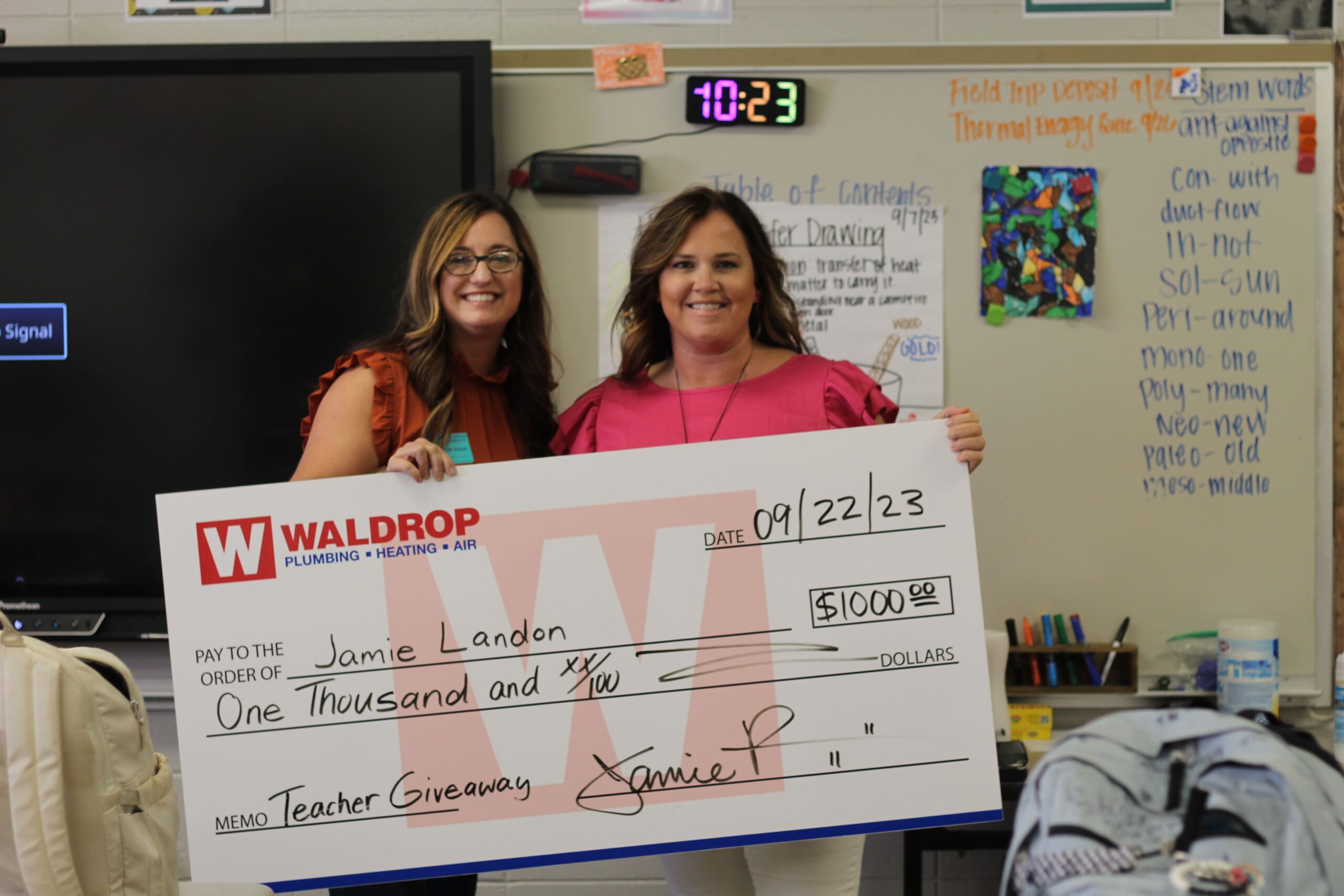 Two smiling women hold a large novelty check in a classroom or school setting, with posters on the wall behind them.