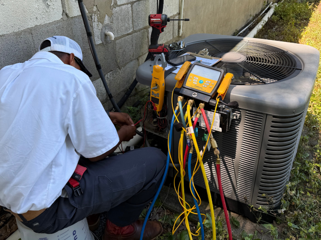 HVAC technician kneeling by an outdoor AC unit, connecting colored refrigerant hoses to gauges and tools set on the unit side.