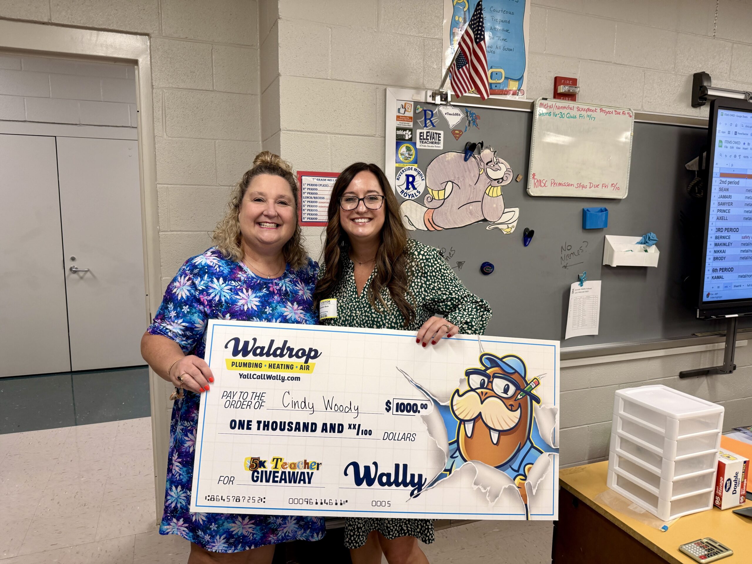 Two smiling women pose with a large novelty check in a classroom setting, proudly displaying the gift amount.