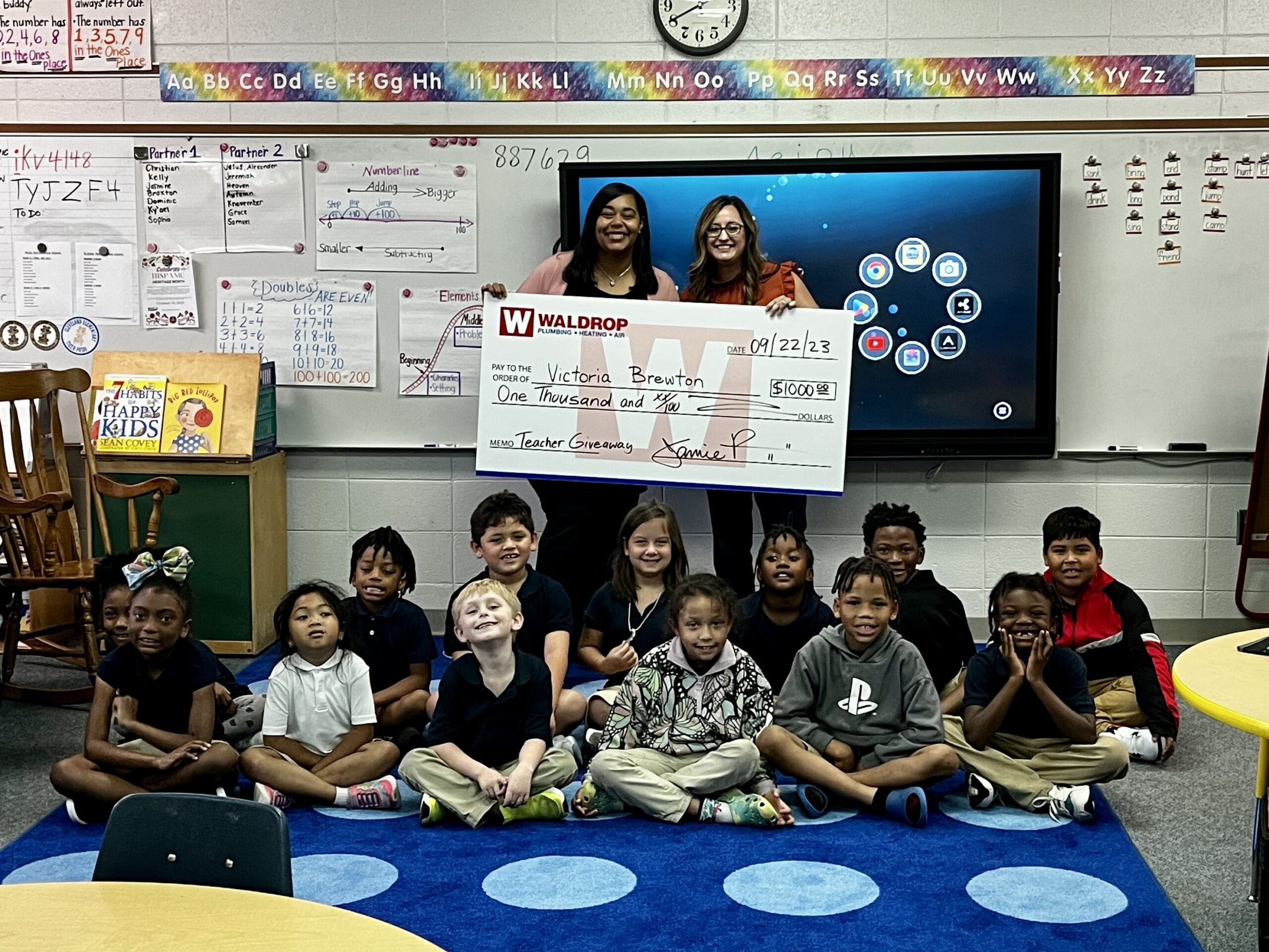 Two teachers grin as they hold a large prize check; students sit cross‑legged on a blue rug in a classroom behind them.