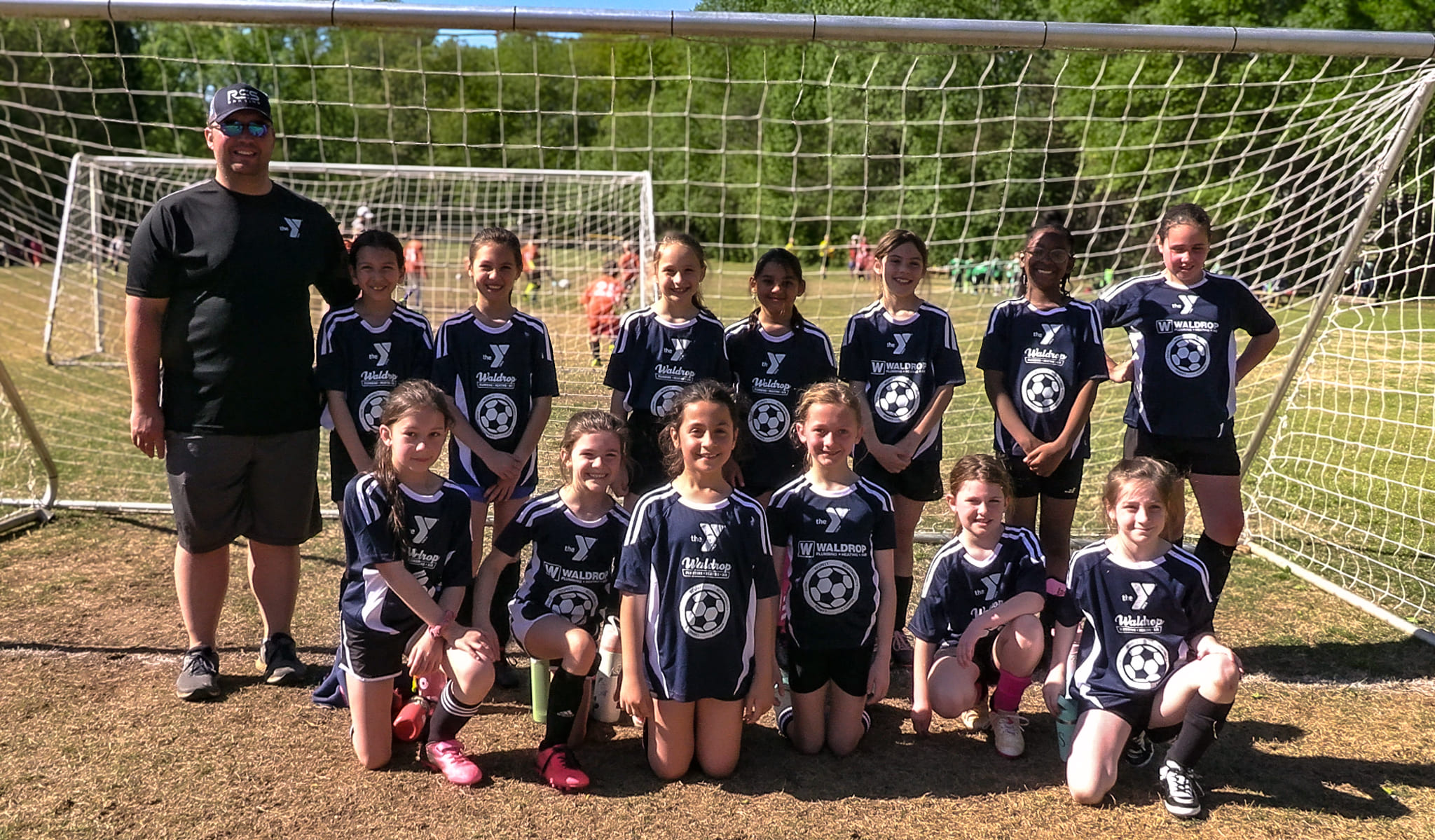 Group of young girls in navy soccer uniforms posing in front of a goal with a coach on the left on a sunny field.
