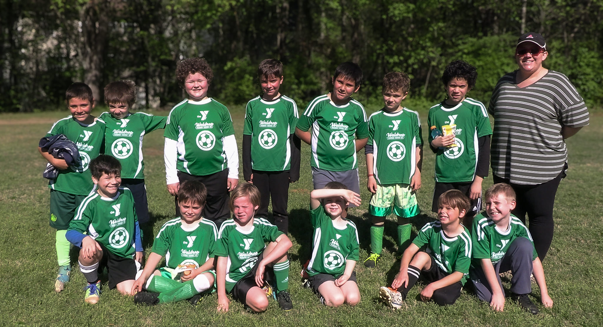 Group of young soccer players in green uniforms posing for a team photo on a sunny grassy field, with a coach standing on the right.