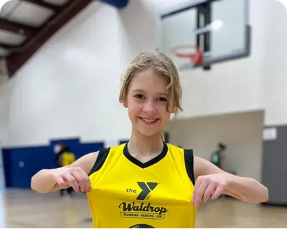 Smiling young girl in a yellow basketball jersey posing and pulling at the shirt in an indoor gym with a basketball hoop in the background.