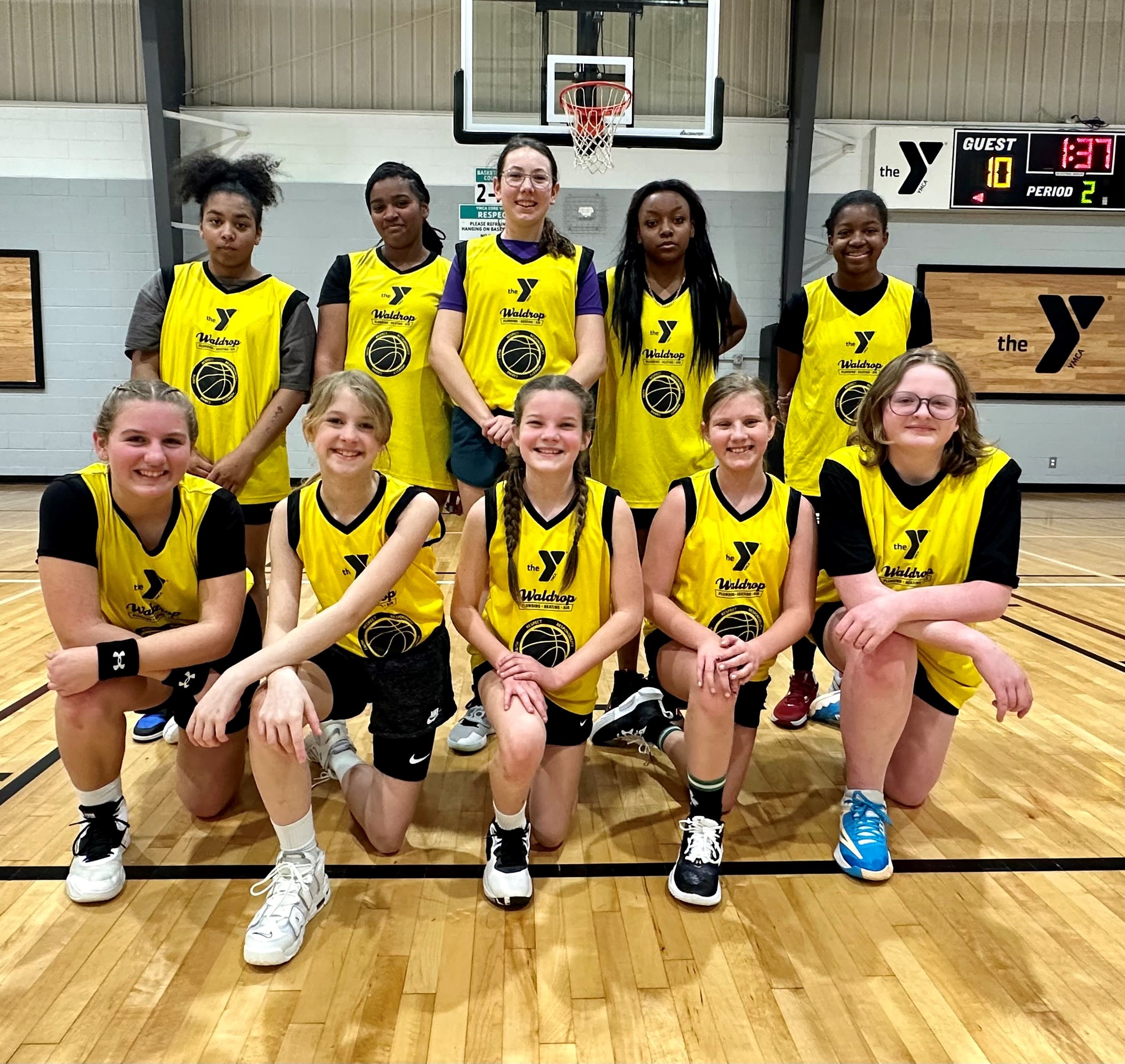 Group of young girls in matching yellow YMCA basketball jerseys posing for a group photo in a gymnasium.