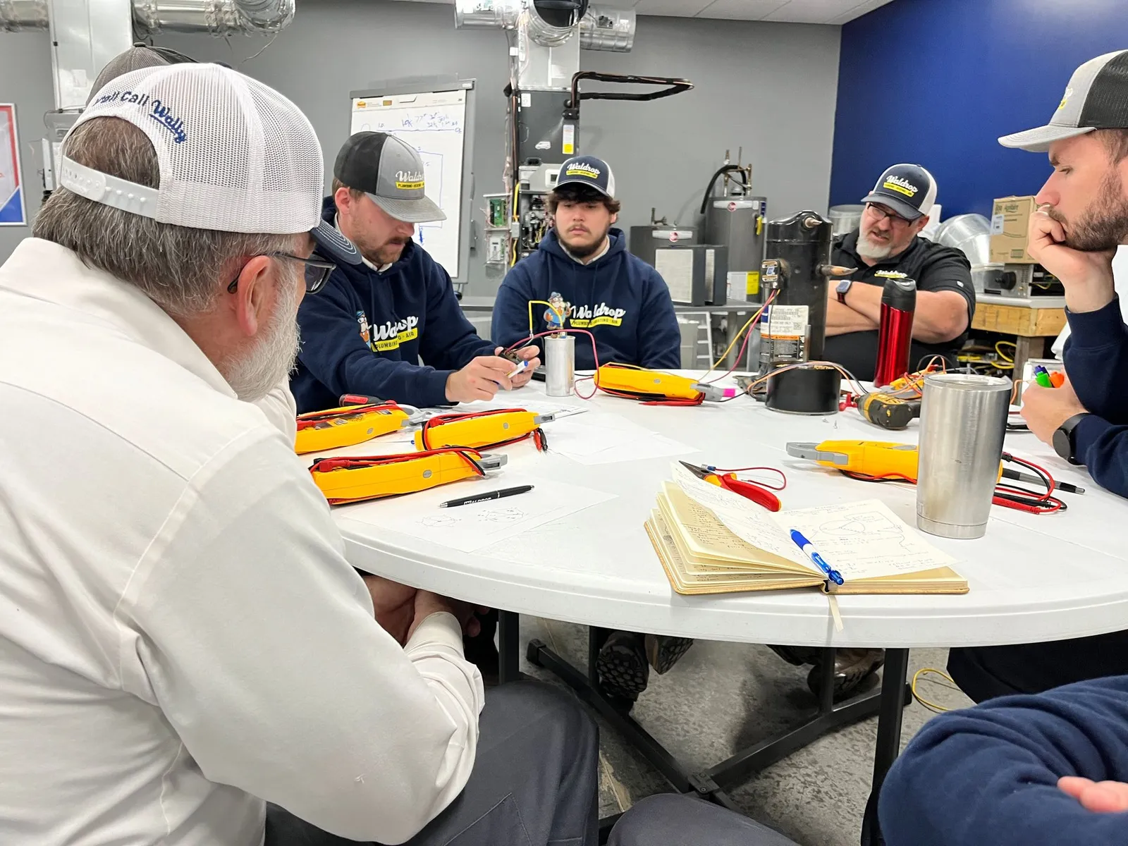 Group of technicians in a workshop gathered around a round table, working with yellow multimeters and notes.