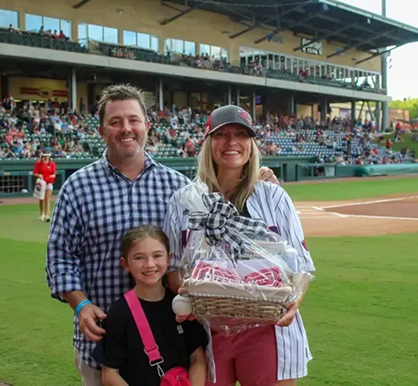 Smiling family of three on a baseball field; woman holding a gift basket as a prize, man beside her, a young girl in front, stadium in background.