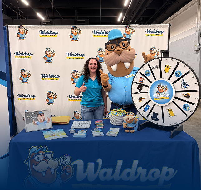 Woman and a bulldog mascot in blue attire pose at a Waldrop booth, giving thumbs up beside a large prize wheel and display table.