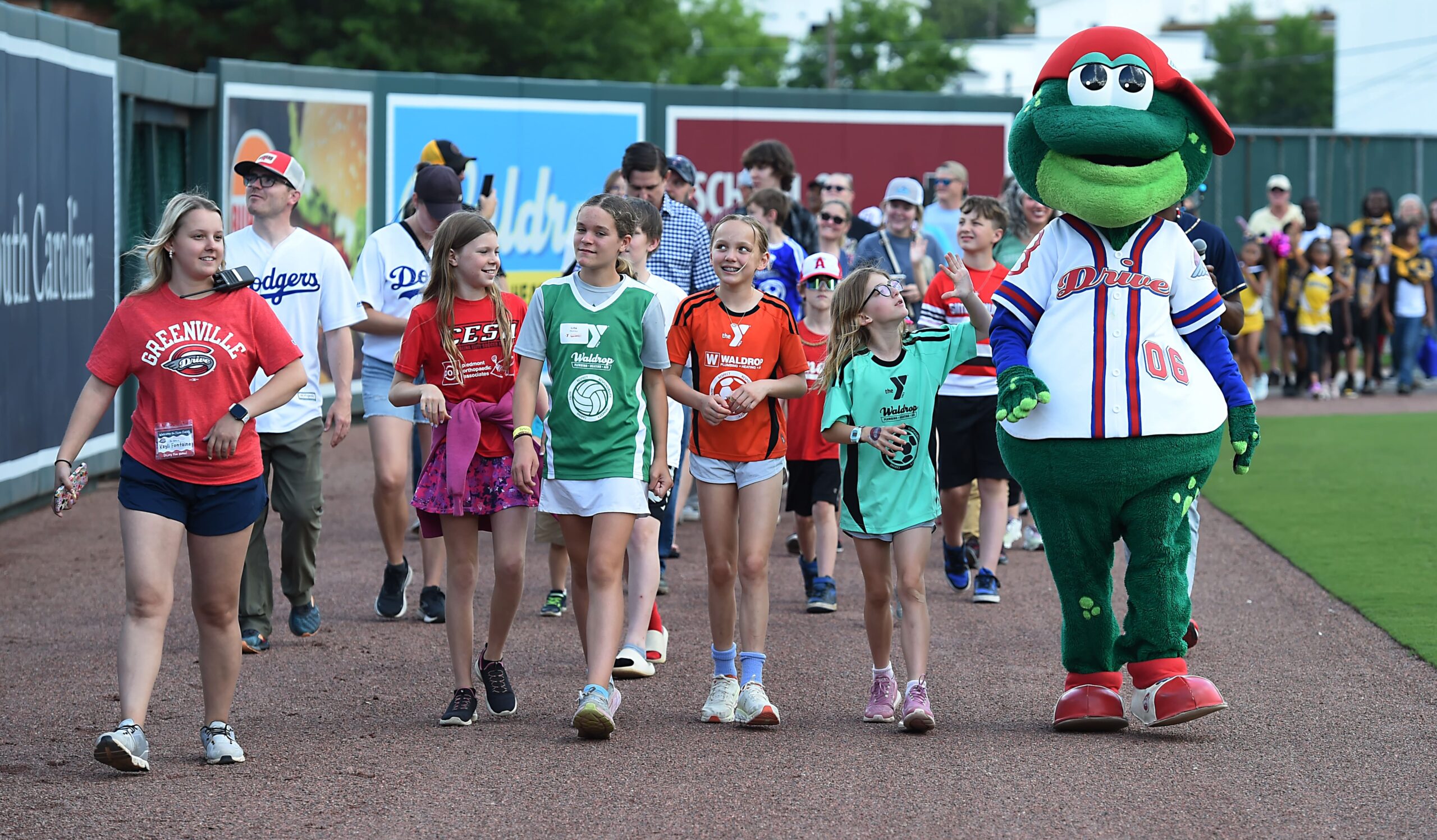 Group of children walking on a track with a large green dinosaur mascot in a red cap and baseball uniform at a sports event onlookers in the background.