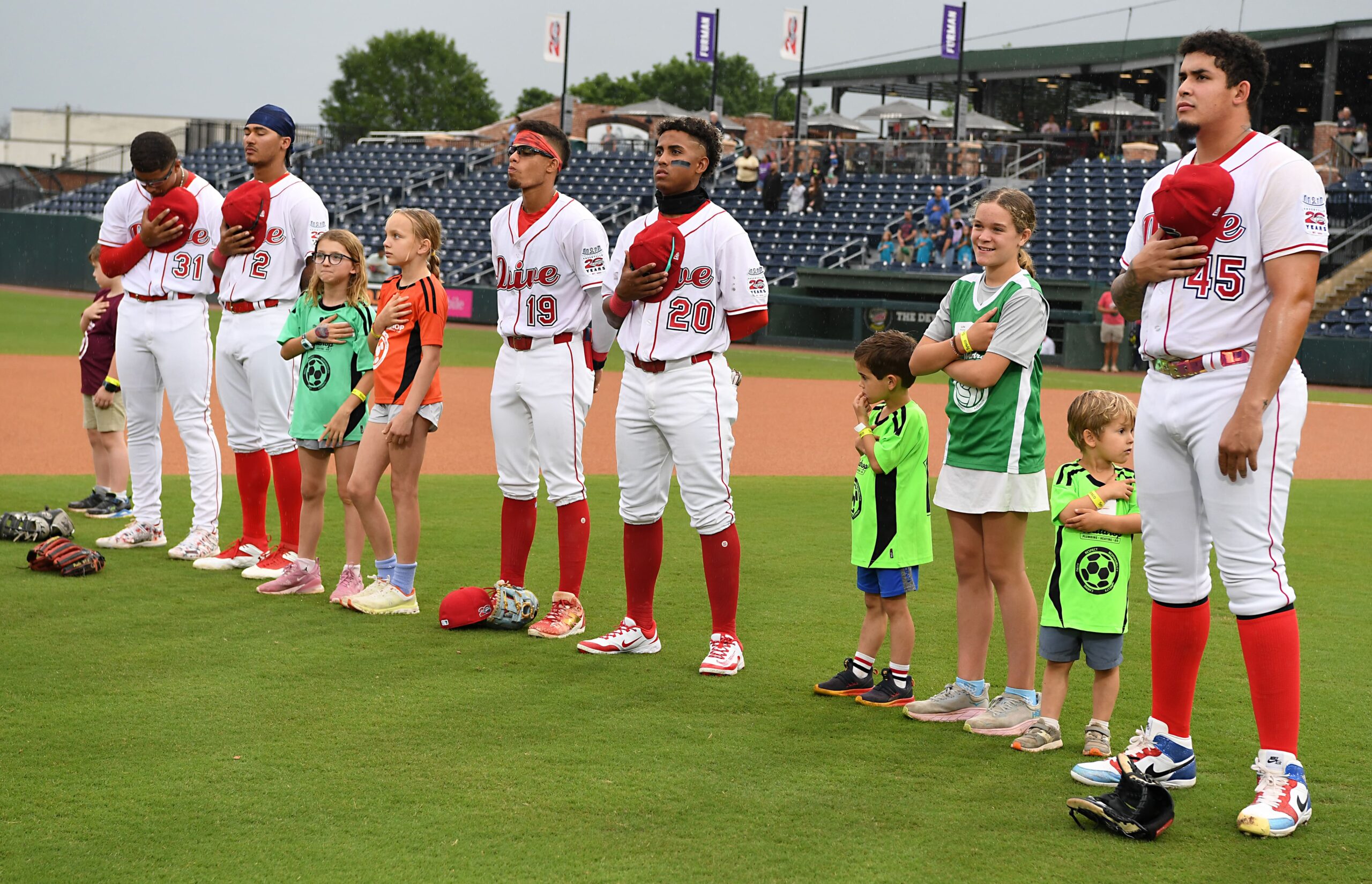 Baseball players in white uniforms with red trim stand with hands over hearts beside smiling kids on the field, stands in the background.