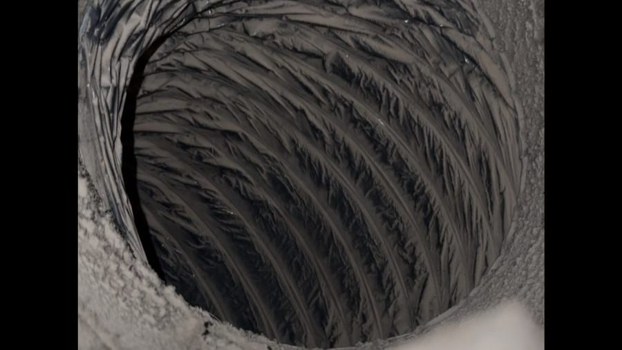 Close-up view of a large ribbed cylindrical tunnel or pipe, looking down into dark, spiraled inner walls surrounded by snow or frost edge.