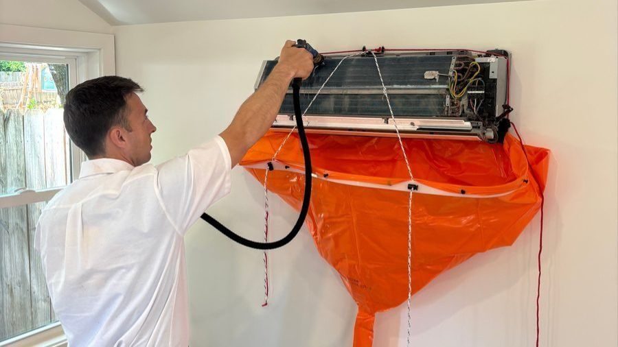 HVAC technician in a white shirt repairs a wall‑mounted air conditioner, with an orange protective bag beneath the unit.