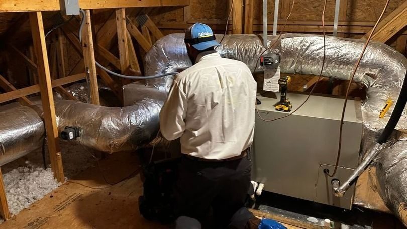 HVAC technician kneels in an attic, working on large insulated ducts near a furnace unit.