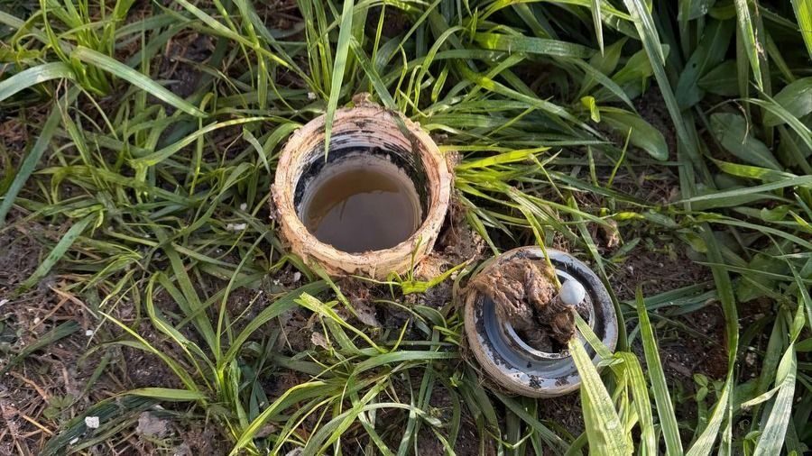 Open irrigation valve exposed in muddy soil among tall grass, with the cap and internal components loosened nearby