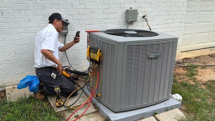 HVAC technician kneels beside an outdoor unit, using gauges and a connected hose while holding a smartphone nearby for readings or notes.