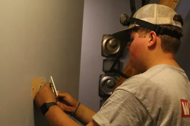 Construction worker wearing a cap and wristwatch, using a tool to attach a small board to a wall.