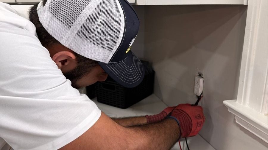Worker in a white shirt and mesh cap kneels at a counter, wiring an electrical outlet with red gloves on.