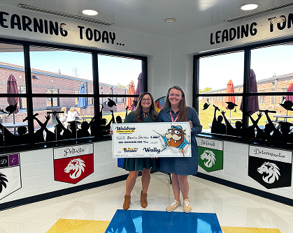 Two smiling students in school uniforms hold a large ceremonial check in a bright hallway during an award event.